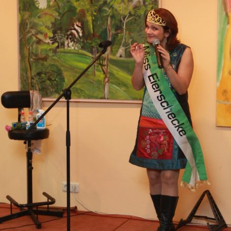 Kathy Leen alias Miss Eierschecke mit Krone und grüner Schärpe steht am Mikrofon, lächelt und gestikuliert.